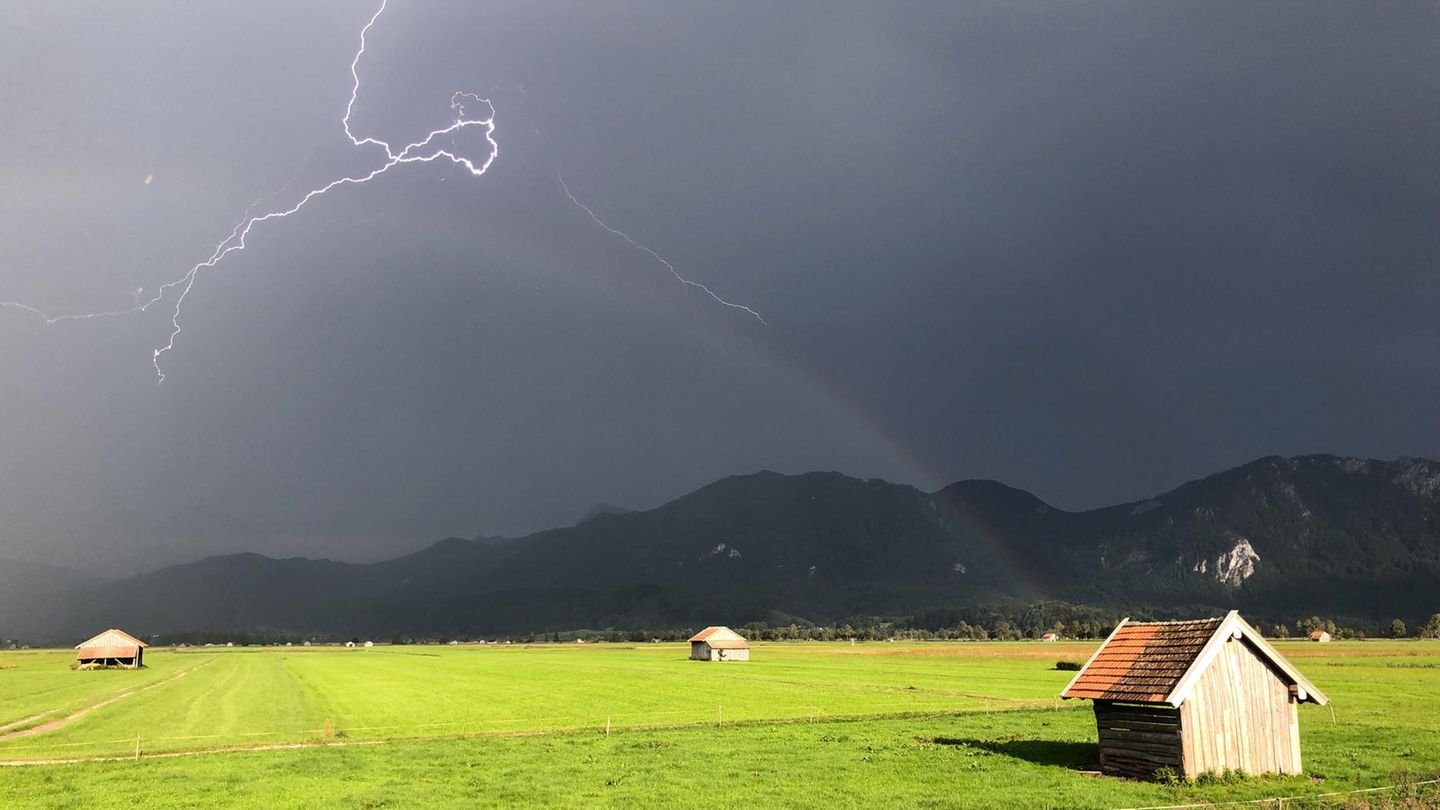 Ein Blitz zuckt über den bayerischen Voralpen, während über einer Weide ein Regenbogen erkennbar