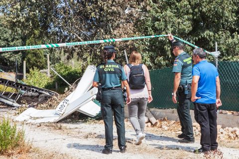 Ein Polizist hält ein Absperrband hoch, damit eine Frau mit Rucksack sich den Trümmern an der Absturzstelle nähern kann
