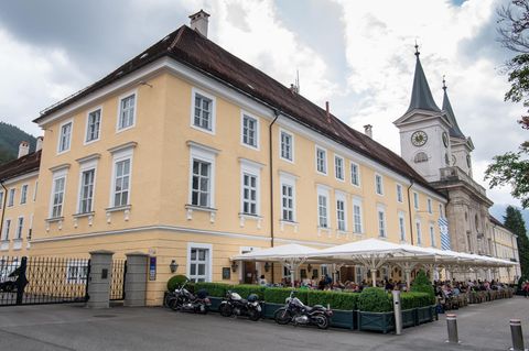 Vor einem dreistöckigen gelben Gebäude stehen weiße Sonnenschirme über den Tischen, an denen Gäste sitzen