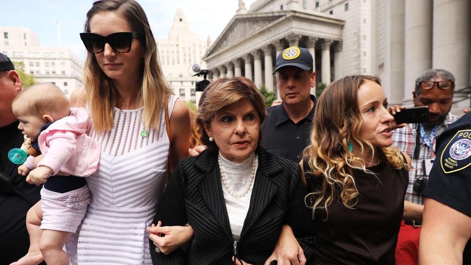 Drei Frauen gehen untergehakt durch eine Menschenmenge vor einem weißen Gerichtsgebäude mit Säulen