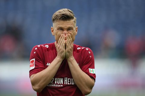Ein Mann mit dunkelblondem Seitenscheitel steht im weinroten Trikot auf einem Fußballplatz und hält sich die Hände vor den Mund