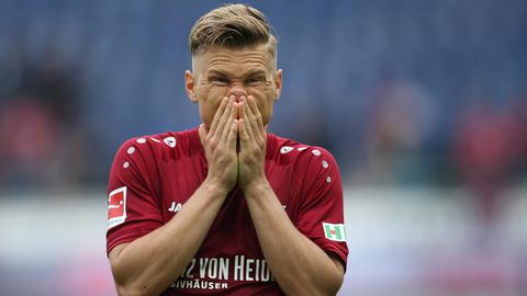 Ein Mann mit dunkelblondem Seitenscheitel steht im weinroten Trikot auf einem Fußballplatz und hält sich die Hände vor den Mund