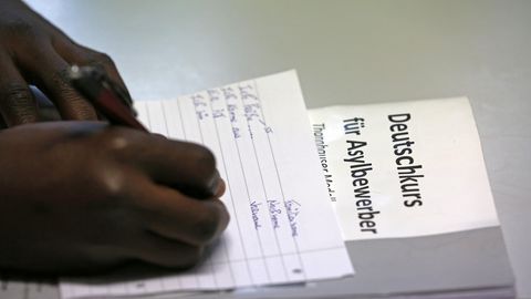 Ein Afrikaner schreibt auf einem linierten Blatt. Darunter liegt ein Papier mit der Überschrift "Deutschkurs für Asylbewerber".