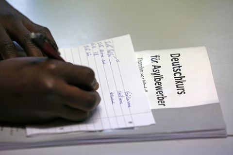 Ein Afrikaner schreibt auf einem linierten Blatt. Darunter liegt ein Papier mit der Überschrift "Deutschkurs für Asylbewerber".