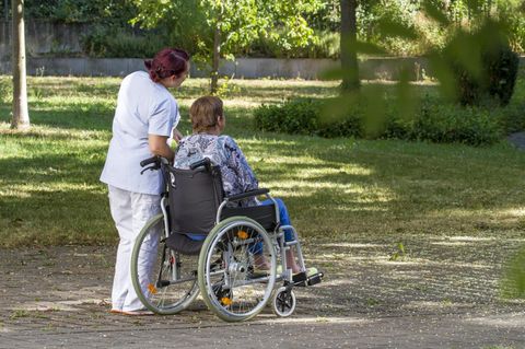 Pflegeheimbewohnerin sitzt im Rollstuhl, Pflegerin bei Spaziergang mit Frau im Rollstuhl.