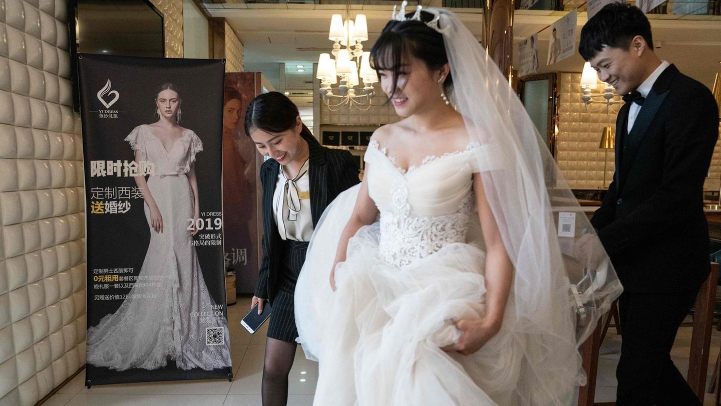 Eine Chinesin in weißem Brautkleid rafft ihren Rock mit den Händen und geht vor ihrem Bräutigam im Anzug her
