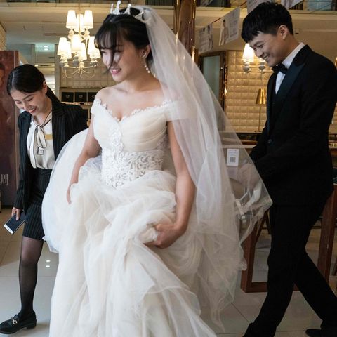 Eine Chinesin in weißem Brautkleid rafft ihren Rock mit den Händen und geht vor ihrem Bräutigam im Anzug her
