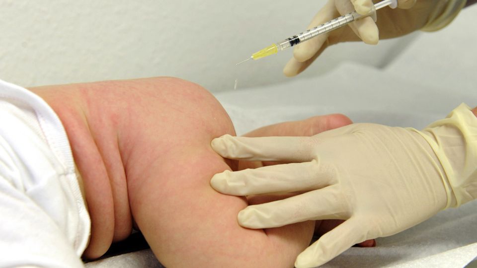 Eine rechte Hand im Gummihandschuh hält eine Spritze, die linke hält den nackten Oberschenkel des auf dem Bauch liegenden Babys
