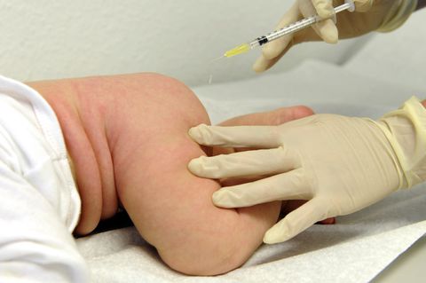 Eine rechte Hand im Gummihandschuh hält eine Spritze, die linke hält den nackten Oberschenkel des auf dem Bauch liegenden Babys