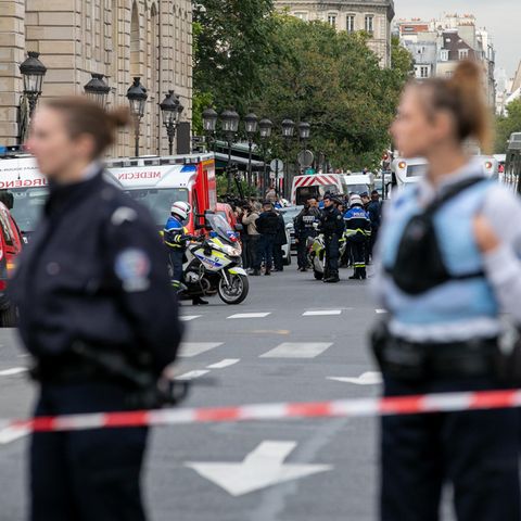 Zwei Polizistinnen stehen in Schutzwesten hinter einem Absperrband, während im Hintergrund Rettungswagen und Polizisten stehen