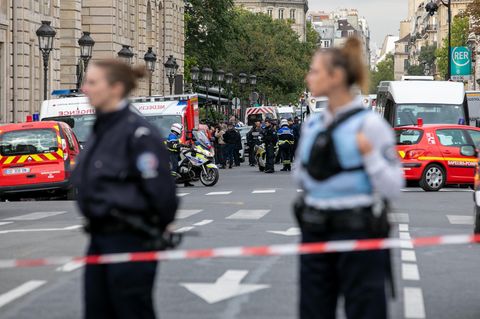 Zwei Polizistinnen stehen in Schutzwesten hinter einem Absperrband, während im Hintergrund Rettungswagen und Polizisten stehen