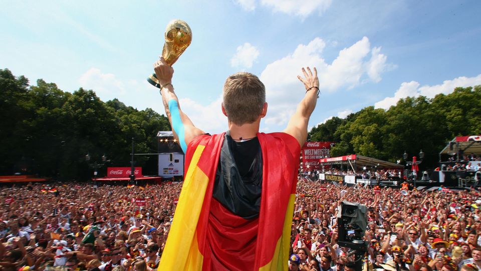 Fußballer Bastian Schweinsteiger hat sich eine Deutschland-Flagge umgebunden und jubelt mit WM-Pokal in der Hand in die Menge