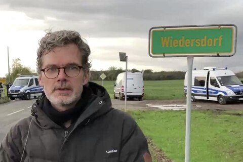 Ein Mann mit Brille, grauen Locken und Bart steht in schwarzer Daunenjacke am Straßenrand. Im Hintergrund Polizeiwagen