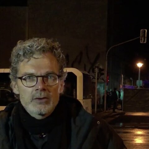 Im Dunkeln steht ein Mann mit Brille und grauen Locken vor einer von der Polizei bewachten Absperrung