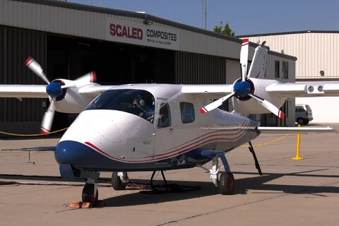 Eine zweimotorige Propellermaschine steht mit rotierenden Propellern vor einem Flugzeughangar