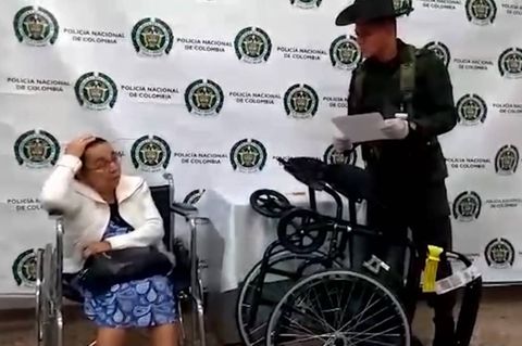 Eine alte Frau sitzt im Rollstuhl, während ein Mann in Tarnanzug und Gummihandschuhen neben ihr steht