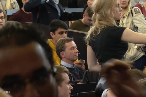 Ein Mann mit braunem Seitenscheitel sitzt in einem vollen Hörsaal zwischen sitzenden und stehenden Studierenden