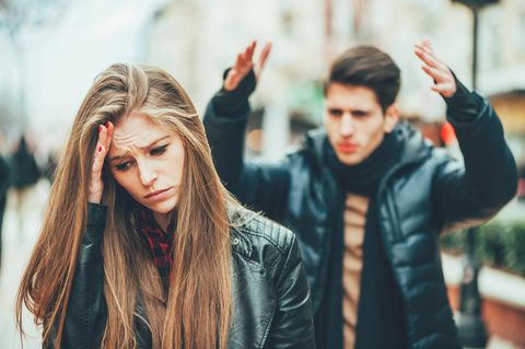 Frau schaut traurig und genervt, während ihr Partner im Hintergrund wütend zu sein scheint