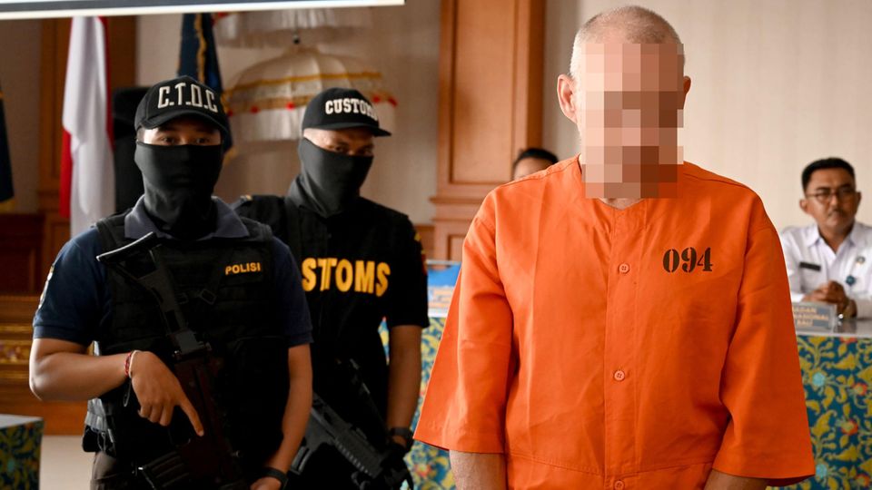 Ein Mann steht in orangenem Hemd vor zwei schwer bewaffneten Polizisten