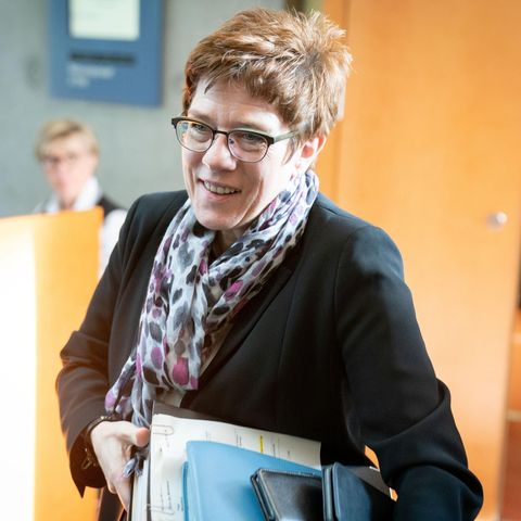 Eine Frau in dunklem Blazer betritt mit blauen Ordnern unter dem linken Arm einen Raum mit hellen Holztüren und -tischen