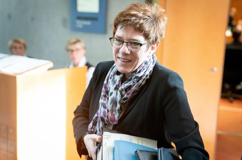 Eine Frau in dunklem Blazer betritt mit blauen Ordnern unter dem linken Arm einen Raum mit hellen Holztüren und -tischen
