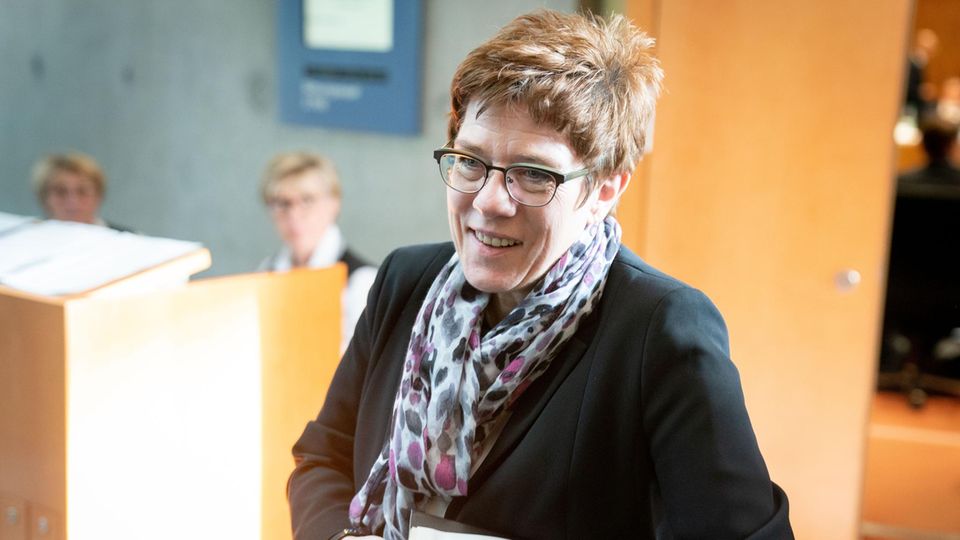 Eine Frau in dunklem Blazer betritt mit blauen Ordnern unter dem linken Arm einen Raum mit hellen Holztüren und -tischen
