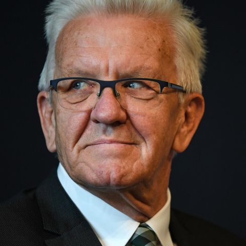 Ein Mann mit weißem Igelschnitt und schwarzem, eckigen Brille schaut mit nach rechts gedrehtem Kopf aus den Augenwinkeln