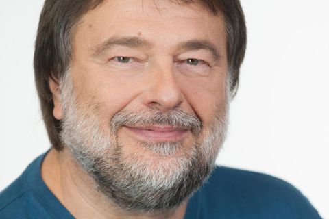 Ein Mann mit braunem Seitenscheitel und grau-weißem Vollbart lächelt in die Kamera