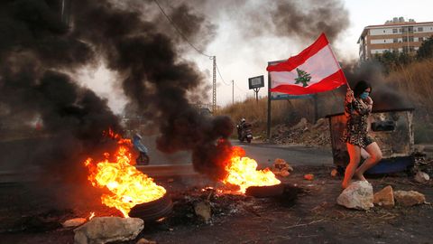 Eine Teilnehmerin einer Demonstration geht mit einer Libanon-Fahne und Mundschutz an brennenden Reifen vorbei
