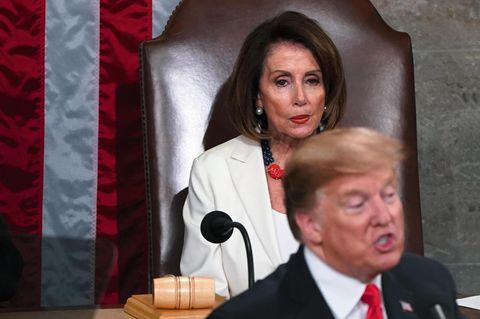 Pelosi, auf einem großen Sessel sitzend, beäugt den vor ihr stehenden Trump