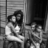 Eine Mutter sitzt mit ihren Kinder vor einem Holzhaus