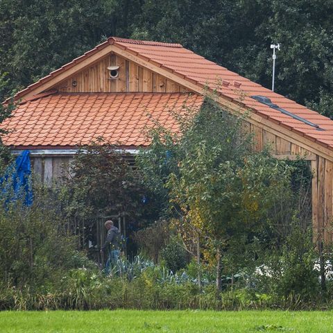 Ein Gebäude mit rotem Ziegeldach und hölzerner Verschalung ist hinter Büschen zu sehen
