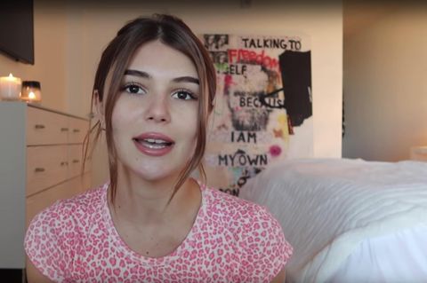 Eine junge Frau mit langen, braunen Haaren spricht in die Kamera. Sie hockt in einem Schlafzimmer und trägt ein rot-weißes Top.