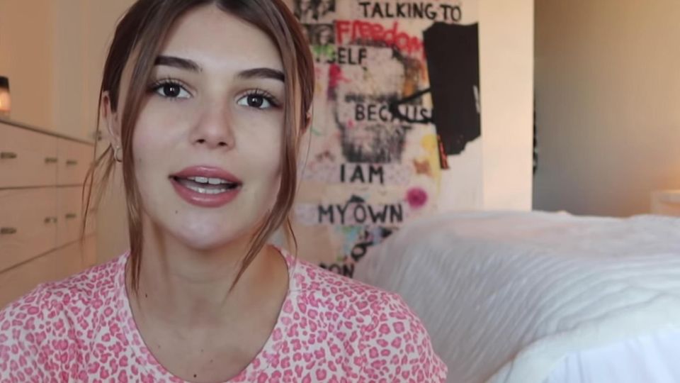 Olivia Jade Eine junge Frau mit langen, braunen Haaren spricht in die Kamera. Sie hockt in einem Schlafzimmer und trägt ein rot-weißes Top.