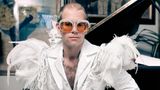 Elton John sitzt vor einem Flügel in weißem Anzug mit Federn auf den Schultern