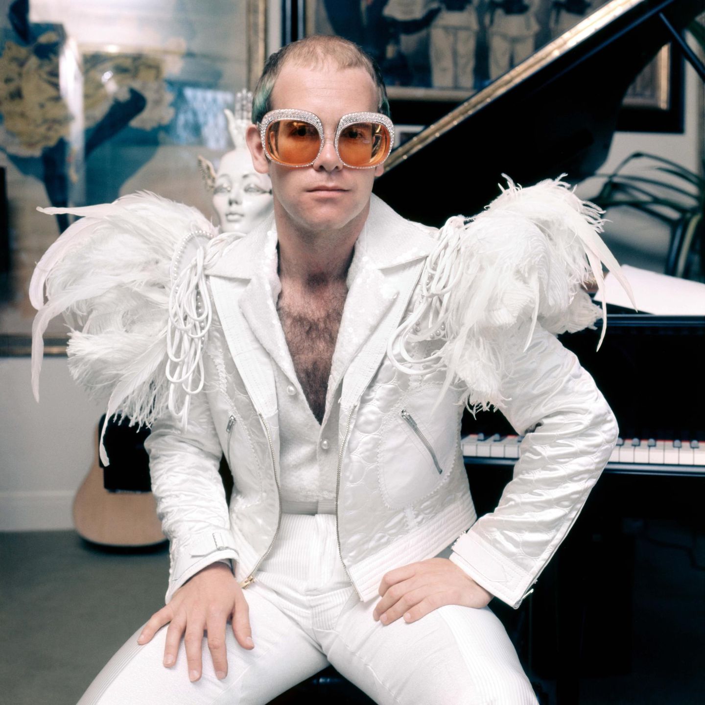 Elton John sitzt vor einem Flügel in weißem Anzug mit Federn auf den Schultern