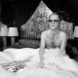 Elton John sitzt in einem Bett und trägt eine riesige Brille