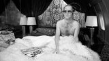 Elton John sitzt in einem Bett und trägt eine riesige Brille