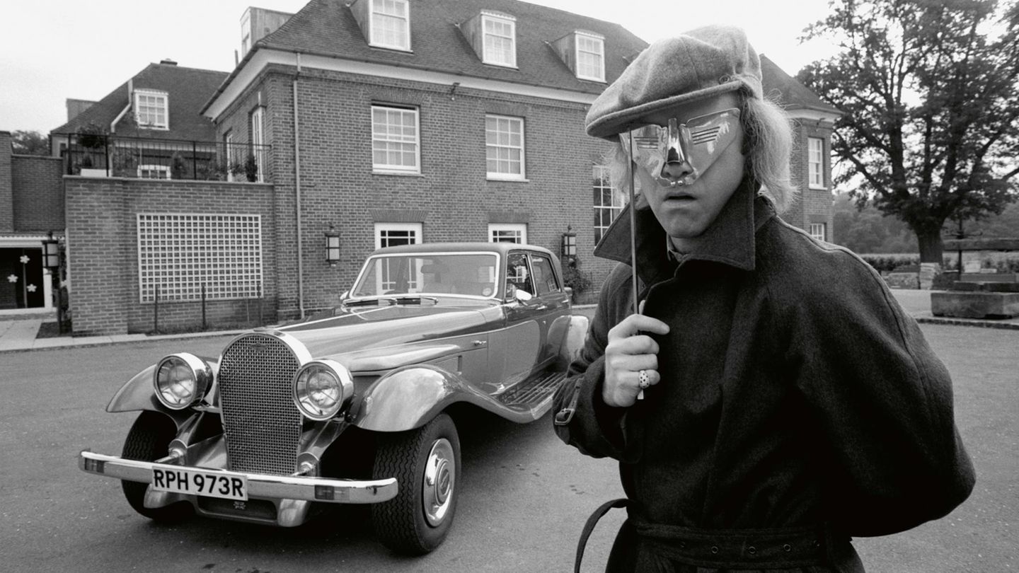 Elton John steht vor einem Oldtimer und hält sich eine silberne Maske vor die Augen