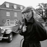 Elton John steht vor einem Oldtimer und hält sich eine silberne Maske vor die Augen