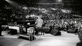Elton John von sitzt mit dem Rücken zur Kamera vor Publikum am Flügel