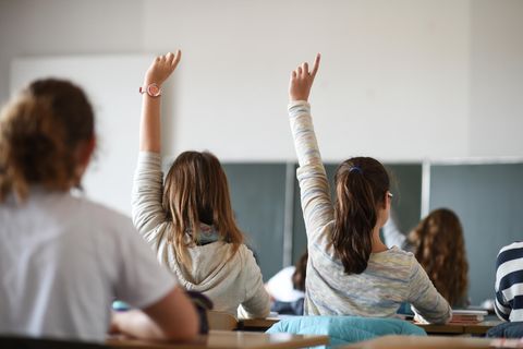 In einem Klassenzimmer sind Schülerinnen von hinten zu sehen. Zwei von ihnen melden sich. Im Hintergrund eine grüne Schultafel