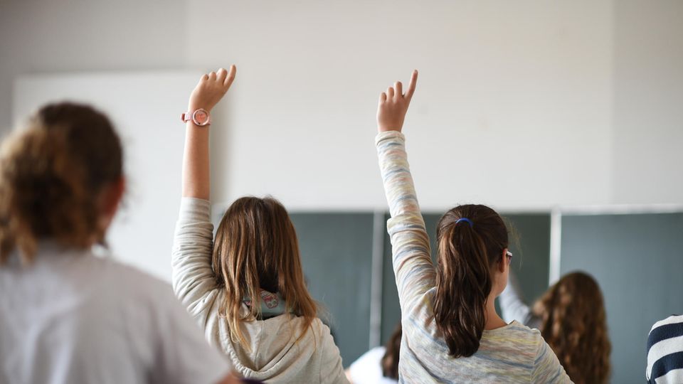 Schülerinnen Klassenzimmer melden In einem Klassenzimmer sind Schülerinnen von hinten zu sehen. Zwei von ihnen melden sich. Im Hintergrund eine grüne Schultafel