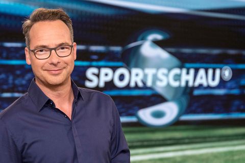 Im dunkelblauen Hemd steht Moderator Matthias Opdenhövel im Fernsehstudio vor dem "Sportschau"-Schriftzug und lächelt