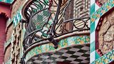 Casa Vicens, 1878–1888, Calle de les Carolines, Barcelona. Prunkvoller kann man sich das Debut eines jungen Architekten kaum mehr vorstellen. Wie ein Märchenschloss aus "Tausendundeiner Nacht" erhebt es sich vor dem Besucher der Calle de les Carolines 24 in Barcelona. Und doch ist es in Wirklichkeit ein recht kleines Haus, auch nicht die Heimat eines Prinzen, sondern das Wohnhaus eines Börsenmaklers.  Straßenseitiger Balkon im Anbau. Orange-gelbe Nelken füllen die farbigen Fliesen, so wie sie Gaudí auf dem noh leeren Grundstück gesehen haben soll.