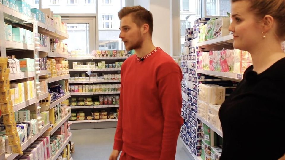 Ein junger Mann in rotem Sweathirt und eine junge Frau in schwarzem Top betrachten ein Supermarktregal voller Periodenprodukte