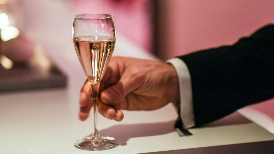 Eine Männerhand greift nach einem Champagnerglas, das auf einem weißen Tisch steht.