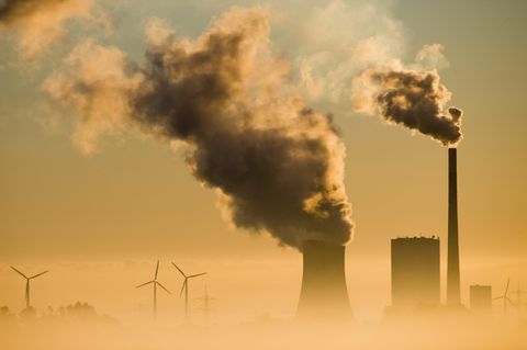 Aus dem Dunst ragen nebeneinander Silhouetten von Kraftwerks-Schornsteinen und Windrädern in den Himmel