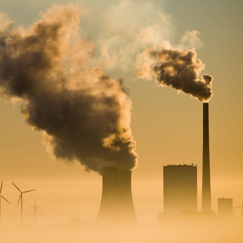 Aus dem Dunst ragen nebeneinander Silhouetten von Kraftwerks-Schornsteinen und Windrädern in den Himmel