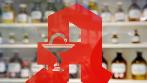 Auf einer Glasscheibe klebt das rote Apotheken-"A", unscharf im Hintergrund braune Apothekerflaschen in einem Regal